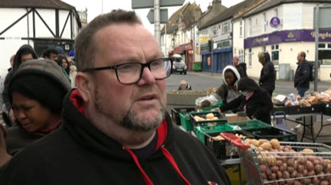 A man with short grey hair and a beard wearing glasses and a black hooded sweatshirt. He is looking off to the right of the camera. behind him are people looking through various crates of vegetables at an outside market beside a road