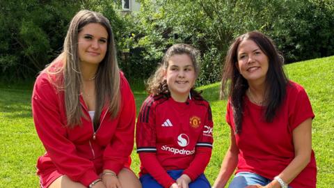 One adult woman, a teenager girls and a young girl wearing red shirts sit on a bench in front of a grassy field. 