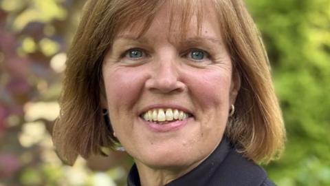 Close up of Rev Jane Atkinson. She is smiling standing outside in front of greenery ad has light brown bobbed hair with a fringe