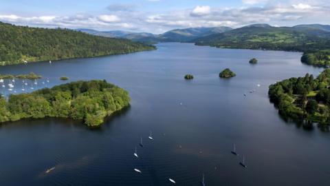 A drone view of Lake Windermere. Boats can be seen o the lake and trees and fields line the lake shoreline. 