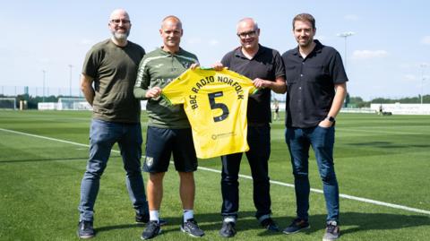Four men standing in a line and looking at the camera. The two men in the middle are holding a yellow football shirt, with the number "5" and "BBC RADIO NORFOLK" printed on the back. They are standing on a football pitch.