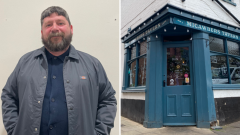 Bun X's Jeff Taylor wears a blue shirt and great coat with exterior view of Micawbers Tavern on right of frame