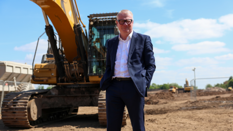 Richard Parker, Mayor of the West Midlands, at the site of the Yardley Brook development in Birmingham with a yellow digger truck behind him.