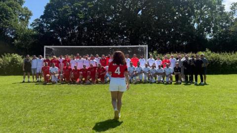 Suzanne Cook walking towards two teams of footballers who are stood on a pitch in front of a football goal.
