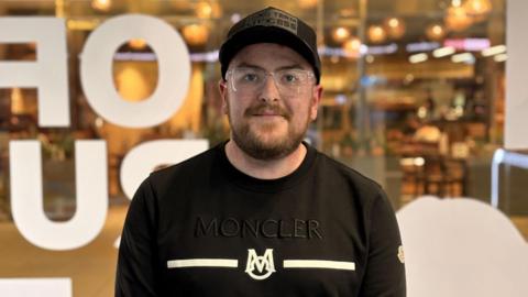 A man wearing a black Moncler sweatshirt, black cap and clear-framed glasses stands with his hands clasped in front of him. He has a short beard. He is standing in front of a glass window with large white letters, with a warmly lit restaurant visible behind him. 