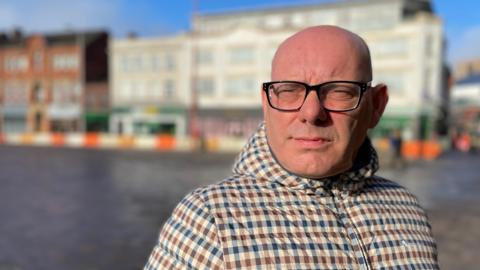 A bald man in glasses stood outside in a town square