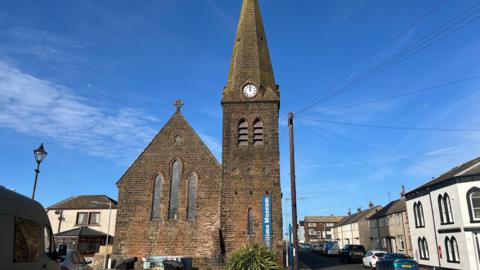 A general view of Christ Church in Maryport. The church is built using red sandstone bricks and has a main building with a pitched roof and three tall, narrow arched windows. Next to it stands a clock tower. There is a blue sign with white lettering with the name Maritime Museum on the clock tower. The building is in the middle of a junction between two residential streets with houses and cars lining them.