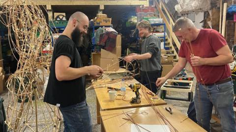 Three men are using masking tape to stick willow canes into a structure. The man on the left has a bald head, bushy beard, glasses and jeans. The man in the middle has a grey jumper rolled up to his sleeves and long fair hair. The man on the right wears a red t-shirt tucked into jeans and a brown belt. He has grey, short hair. All three men are smiling. They are working either side of a wooden workbench, covered in tools and materials. They are all inside a large workshop with numerous boxes of tools and supplies on shelves.