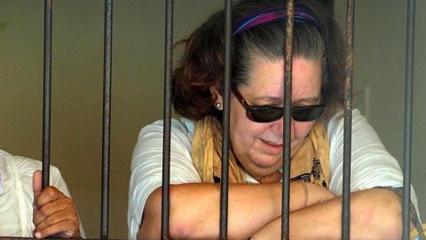 Lindsay, who has dark hair pushed back by a purple headband, wears a white top, a yellow scarf and dark sunglasses. She is behind bars and looking down towards her crossed arms. 