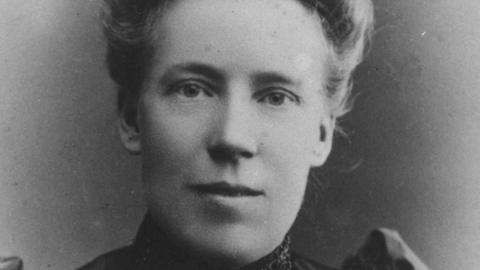 A black and white close up head shot of a woman. Her hair is pinned up and she is looking at the camera. 