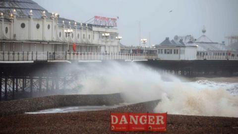 Brighton Pier and its white buildings, extending into the sea. The water is rough, with large white waves. There is a danger sign on the pebble beach.