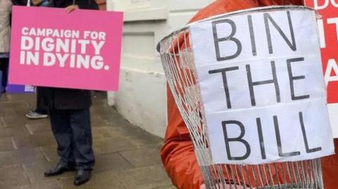 A pink sign which says campaign for Dignity in Dying is held up alongside a metal bin which has a white sign saying bin the bill attached to it.