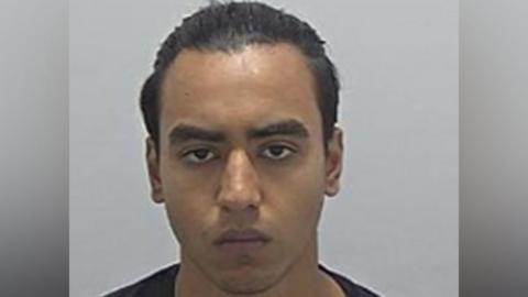 Nazim Uddin's police custody photo, looking straight at the camera. He has short, slicked back, dark hair. He is wearing a dark top. 