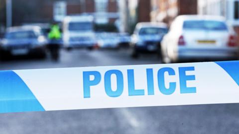 A close up shot of blue and white police tape. It is striped with "police" written on it in blue capitals. In the background there are a number of cars on a residential street, although it is blurred quite heavily.