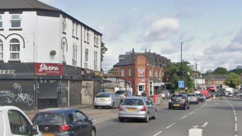 The junction of Ladypool Road and Stratford Road in Birmingham. The daytime photograph shows cars queueing in the city and road markings for a junction, with a bollard just visible. There are shops and other businesses along the road. 