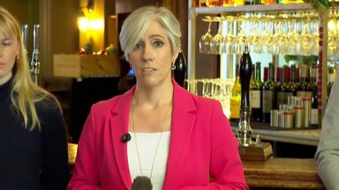 Daisy Cooper speaking in pub. Behind her a pumps and bottles, and a bar. She is wearing a bright pink jacket and white t-shirt and staring at the camera as she speaks.