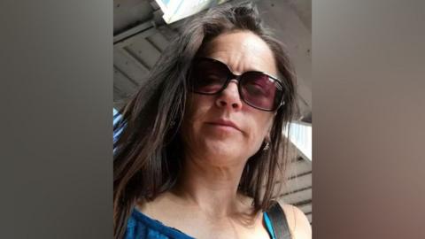 Joanne Sheen has long, dark brown hair and wears sunglasses and a lacey blue, sleeveless top.