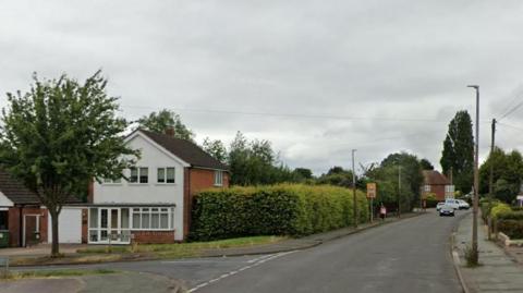 A residential road lined with houses, bushes and trees. Cars can be seen in the background.