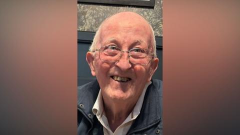 Dessie Sheridan, an elderly man with glasses, smiles to the camera. He is bald on top and has grey hair at the sides.