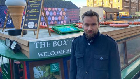 Leo stands bedside the narrowboat with a serious expression on his face. He has brown hair and a beard, and is wearing a smart button up coat with large pockets. The green boat is behind him. 