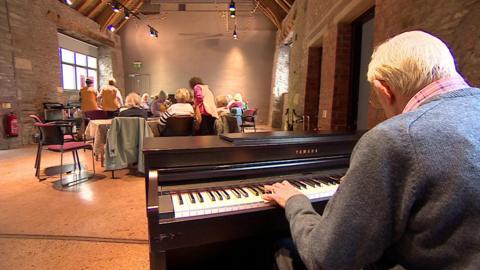 Image of a man, photographed from behind, sitting down at an upright piano.  He is wearing a blue jumper and a red checked shirt and has white hair and is wearing glasses.  In the background can be seen a number of people sitting with their backs to him at a number of small tables.