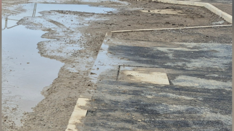 A pavement on the right and road on the left which has not yet been covered by tarmac. There is a huge large puddle in the road and the rest is covered by mud. The mud has been dragged onto the pavement.