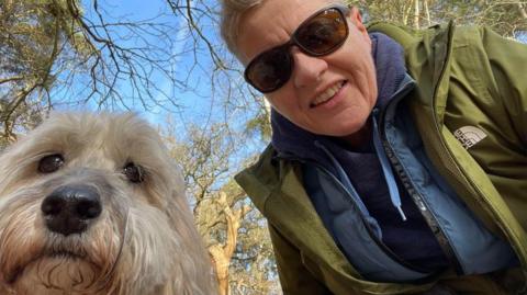 Murphy a golden brown cockapoo and his owner Joanne are close up to the camera in a selfie in the woods. Joanne wears sunglasses and layers of outdoor clothing, including a khaki green North Face anorak.