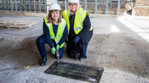 Councillor Elizabeth Scott and councillor Amanda Hopgood they are both kneeling down and wearing hard hats and hi-vis jackets. Ms Scott is holding a drill which has been placed on the metal plate. They are inside the DLI Museum building site.
