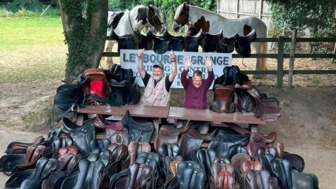 Two men sitting behind a row of saddles. There are two horses behind them.