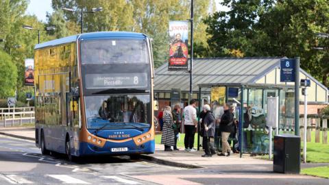 A double-decker bus marked "York Park & Ride" is parked at a sheltered bus stop. The bus displays route number 8 with the destination "Piccadilly" on its front. Several people are gathered nearby—some waiting, others boarding. 