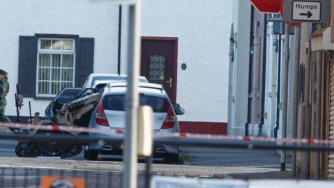 A police officer and cars visible on a residential street. There is police tape across the top of the street. 