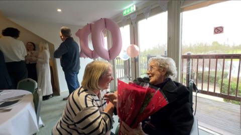 The BBC's Sophie Law crouches down to present 100-year-old Dorothy Howard with a bouquet of red roses. Pink balloons of the number '100' can be seen in the background.