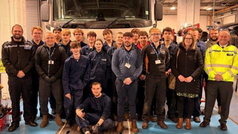 A large group shot showing mechanics students in blue overalls, teaching staff from Carlisle College and police officers in uniform. They are all standing in front of a truck and posing for the picture.