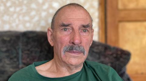Nigel Stevens-Astell sitting on a sofa, wearing a green T-shirt. He has short grey hair and a grey moustache. In the background is a wooden door and patterned wallpaper.