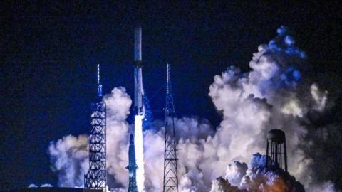 A space rocket launches in a trail of blue flames and light, leaving smoke as it lifts off.