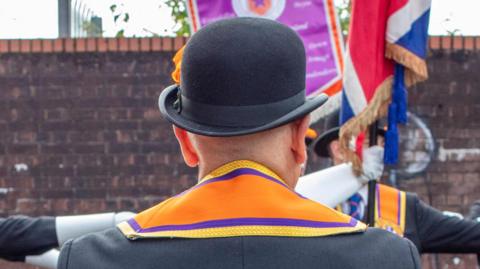 A head and shoulders images taken from behind of a man in a black bowler hat.
He is wearing a dark suit and has an orange sash around his neck.
There is a brick wall in  the background. In front of it is another man in a dark suit but his suit is obscured by a union flag.
