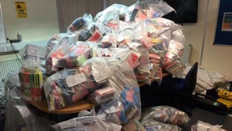 A large number of see-through plastic bags in a room and on a table. They all contain colourful vape boxes.