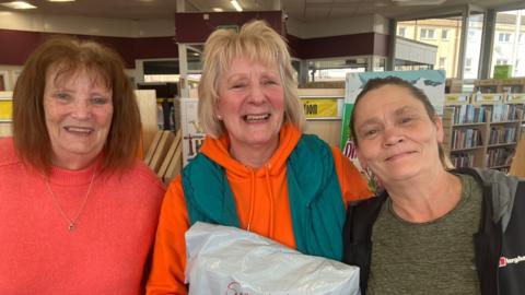 Three women stand together smiling in front of bookshelves in a library