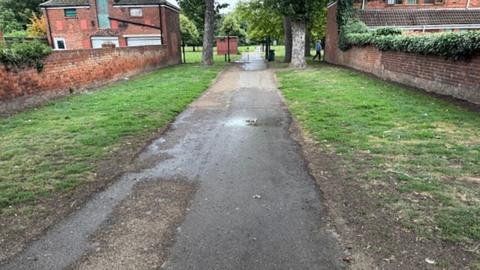A footpath in East Park the tarmac is uneven and has patched up holes. The path is flanked by grass and two houses on either side and a metal gate at the the end of the pathway