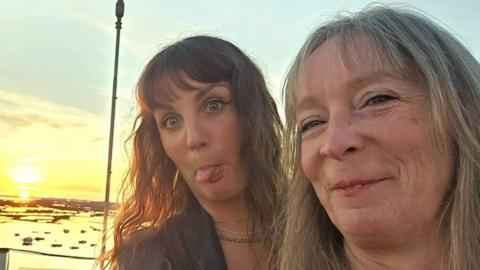 A mother and daughter taking a selfie as the sun sets on a beach. The daughter is sticking her tongue out and the mother smiles at the camera