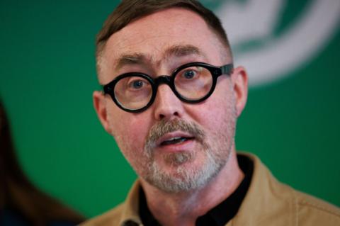 A man, with brown hair and stubble. He is wearing round, black glasses. The background of the image is green. 