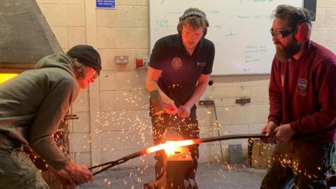 Rowan Pickford, Kyle Priestman and Rowan Taylor are demonstrating forge welding techniques at the National School of Blacksmithing. Kyle Priestman is hitting a molten piece of metal with a hammer and orange sparks are flying everywhere.