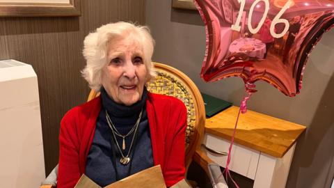 A woman with white hair in a red cardigan and black polo neck. She is wearing gold necklaces and has a helium balloon with 106 on it.