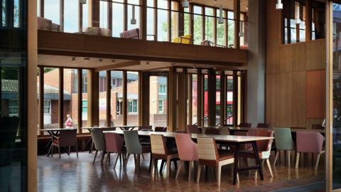 Looking in through large open glass doors to a double-height timber-walled communal room with a large table and chairs of different colours
