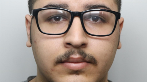 A custody picture of a young man with a moustache wearing glasses.
