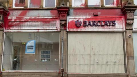The outside of a bank building, which has a white sign with the word 'Barclays' in blue lettering. Red paint has been splashed over the signage and cracks are visible i the window