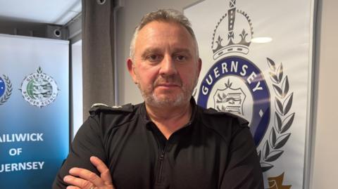 Guernsey Police's Chief Officer Damian Kitchen stands in front of a Guernsey Police sign with his arms crossed against his chest. He is wearing a black police uniform. He has short grey hair and a trimmed grey beard.