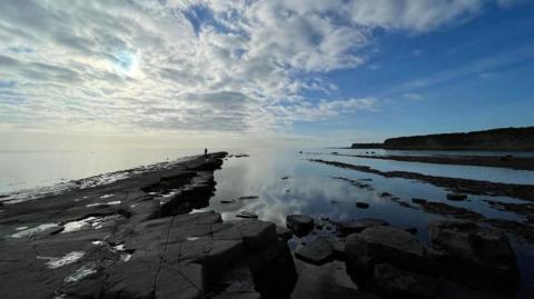 Dark mudstones half submerged in the sea, with a thin stretch of the rocks heading out further than the rest and a person can be seen walking along them.