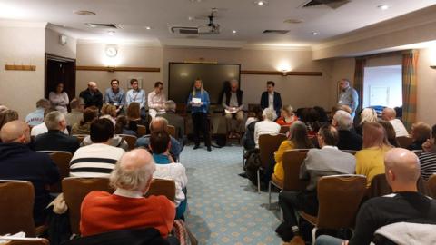 Lots of people sat in a meeting room looking at a blonde haired women in a blue jumper and black trousers holding papers.