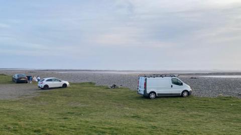 Three vehicles parked on some grass in front of a pebbled beach. The closest is a white van, with a white car in the middle and black car in the distance with people around it. The sky is calm and the sea can be seen in the distance.